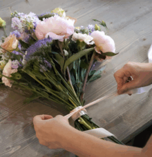 Händer binder band runt en bukett med blandade blommor på ett träbord.